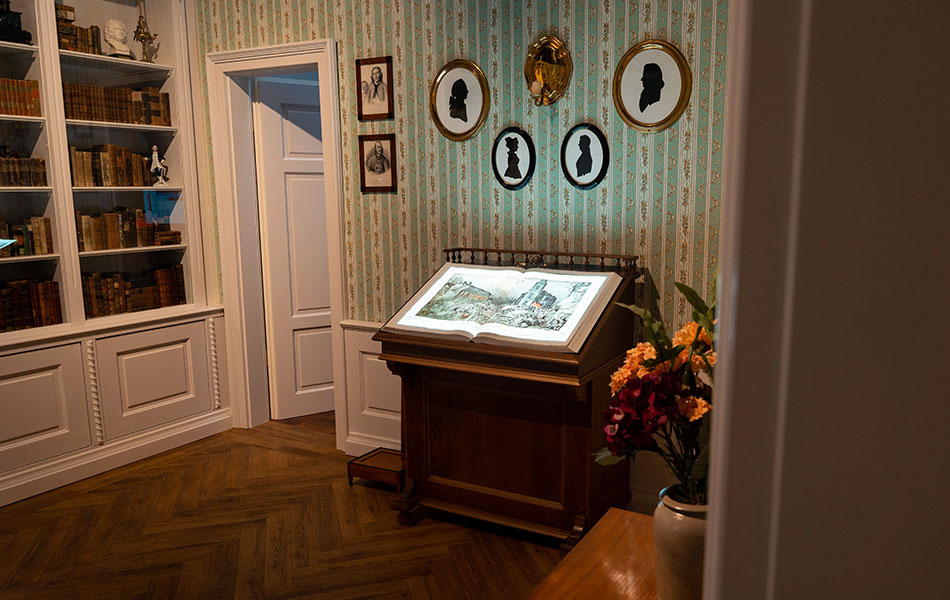Historisch dekorierter Raum mit einer Vitrine an der Wand auf der ein Screen in Form eines Buches liegt.