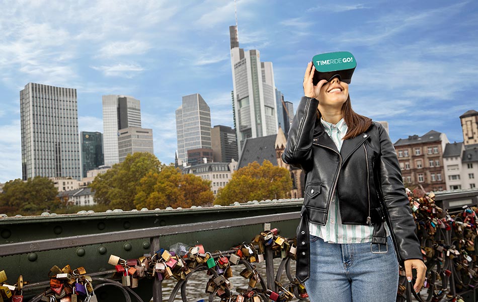 Frau mit VR-Brille vor moderner Frankfurter Skyline auf Brücke. Vorhängeschlösser hängen am Brückengeländer. TIMERIDE GO!