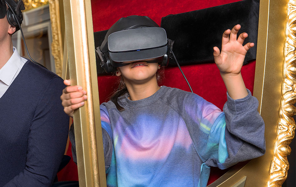 Kind in einer goldenen Kutsche mit buntem Sweater und VR-Brille greift nach etwas.